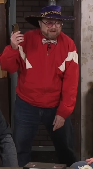 Rich Evans wearing rose-tinted glasses, a red track suit, a bowtie, and a pimp hat.