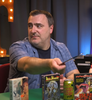 Mike sitting at the Best of the Worst Table, tapping ''Blood Freak'' with a bottle opener. Near his seat is a can of Controlled Chaos.